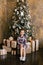 A five-year-old girl in a plaid dress sits near a Christmas tree on new year`s gifts and smiles