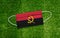 Flag Angola placed on a medical mask lies on the green grass. Covid-19 pandemic concept