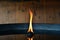 The flame of an oil lamp is seen against a wooden wall, which is reflected in the oil