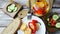 Flat lay of marinated cheese in olive oil with capsicum in glass jar, fork, plate tomato bread and cucumber on wooden background