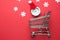 Flat lay of winter holiday shopping made of shopping trolley , small clock with santa hat and white snowflakes isolated on red