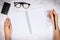 Flat lay of woman hand writing in a blank white page notebook on white marble desk, top view. glasses and mobile smart phone