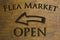 Flea Market Open sign set on a wood background and a left facing arrow. Brown 3D text against wood grain background.