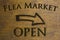 Flea Market Open sign set on a wood background and a right facing arrow. Brown 3D text against wood grain background.