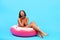 Flirty black lady in bikini sitting in inflatable ring, blowing air kiss at camera on blue studio background