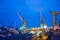 Floating dock in shipyard at dusk, vessels undergoing repair, industrial cranes tower above. Marine maintenance