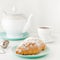 Flying food concept. Powdered sugar falling on freshly baked croissant on white breakfast table with teapot and tea.