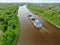 Flying over a large cargo ship with open holds sailing down a calm river surrounded by forest. Carriage of goods by river.