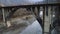 Flying under rusty bridge crossing the narrow river with stony bottom surrounded by mountains and forest. Shot. Aerial