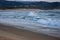 Foaming ocean waves breaking on sandy shore in evening dusk. Surf on beach with village and hills on background.