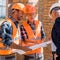 Focus of constructors near mature businessman in helmet looking at blueprint