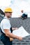 Focus of handsome builder in helmet holding blueprint near colleague repairing roof