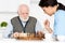 Focus on retired man sitting on kitchen, and playing in chess with nurse