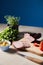 Focus of tasty ham on cutting board with knife, parsley, cherry tomatoes, olives on wooden table on blue background