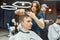 Focused barber girl holding scissors and comb in hands and making haircut for her client. Young guy visiting barbershop