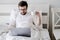 Focused bearded man, with a cup of coffee in his hand, works on a laptop, sitting in bed, under natural light. Modern