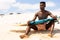 Focused biracial man holding surfboard sitting on sunny beach looking away