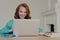 Focused cheerful redhead successful businesswoman does research, makes business project, sits in front of laptop computer at home