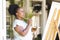 Focused happy senior african american woman holding paint palette painting canvas on easel at home