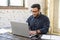 Focused hindu freelancer guy wearing eyeglasses using laptop in contemporary office space