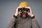 Focused mature man in warm sweater and knitted hat looking at camera through binoculars