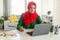 Focused muslim woman in hijab using laptop and calculating budget, sitting at table in living room interior