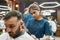 Focused professional barber girl working with hair clipper, making modern haircut for a young handsome man visiting