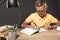 focused schoolboy doing homework in textbook at table with colour pencils lamp and stack of books on grey
