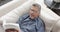 Focused senior biracial man in glasses lying on couch reading book at home, slow motion