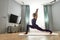 Focused tattooed young woman in bodysuit looking away while exercising, doing High Lunge yoga pose on a mat in living