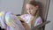 Focused teenager girl in pajama reading book in chair. Young schoolgirl in home clothes reading book sitting in chair in