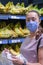 Focused woman in face mask choosing fruits, taking bananas from shelves in grocery store. Customer in supermarket. Side