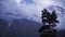 Fog clouds above mountains and forest. Clip. Mysterious landscape with snowy mountain peak and heavy sky with a pine