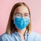 Foggy glasses wearing on woman. Woman in medical protective face mask and eyeglasses wipes blurred foggy misted glasses on pink