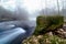 Foggy winter landscape, river with water falling down a waterfall, rocks and bare trees. Long exposure photo