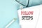 FOLLOW STEPS text on a sticky on notebook with pen and glasses , blue background