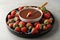 Fondue fork with strawberry in bowl of melted chocolate surrounded by different berries on grey table