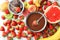 Fondue fork with strawberry in bowl of melted chocolate surrounded by other fruits on white marble table, flat lay