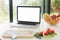 Food blog. Laptop with blank white screen and vegetables, knife and fruits on countertop in modern kitchen with big windows, copy