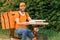 Food delivery man in orange uniform sitting on a moped with a food delivery bag and holding boxes of pizza