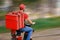 A food delivery man in a red uniform rides a moped with a food delivery bag.