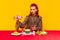 Food pop art photography. Beautiful stylish redhead woman sitting at table against yellow background and having dinner