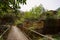 Footbridge over weedy river before deserted 1970s` buildings of factory