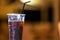 In Foreground In Focus Is Plastic Cup With Dark Cold Drink And Black Straw, In Background Out Of Focus Is Empty Beach