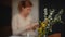 In the foreground, a spring bouquet in a glass vase stands on a table indoors. A pregnant woman, out of focus, sits and