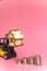 A forklift truck holds a model of a house on a pink background on a platform. The concept of buying real estate