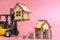 A forklift truck holds a model of a house on a pink background on a platform. The concept of buying real estate