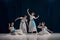 Four ballerinas dressed in white dresses with blue sashes pose gracefully against dark backdrop on theatrical stage.