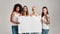 Four diverse women looking at camera while holding, standing with blank banner in their hands isolated over grey