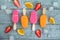 Four pink and yellow ice creams on a stick on a gray wooden background next to juicy pieces of fruit and ice.